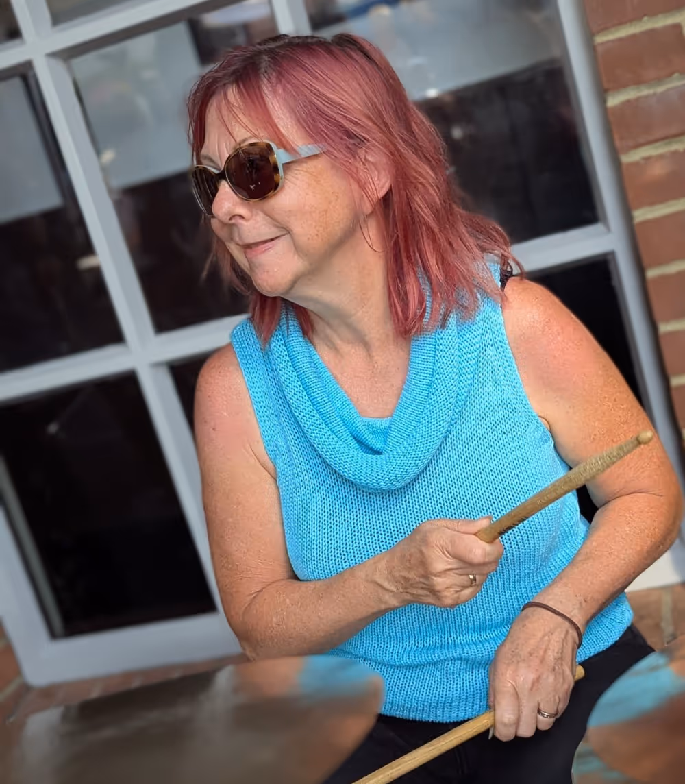 A woman with purple hair wearing a turquoise top and playing the drums.