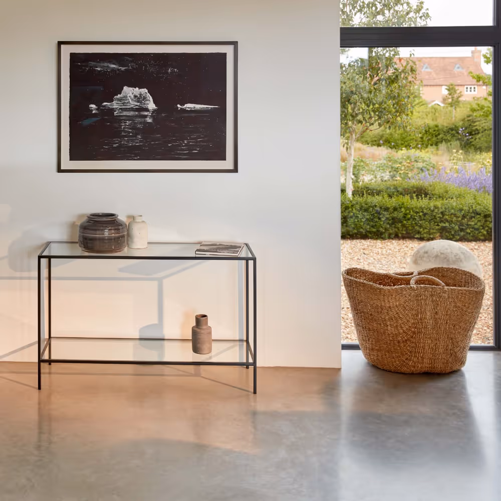 A modern room with white walls a metal and glass console table, a wicker basket and a floor to ceiling looking out to a lush garden.