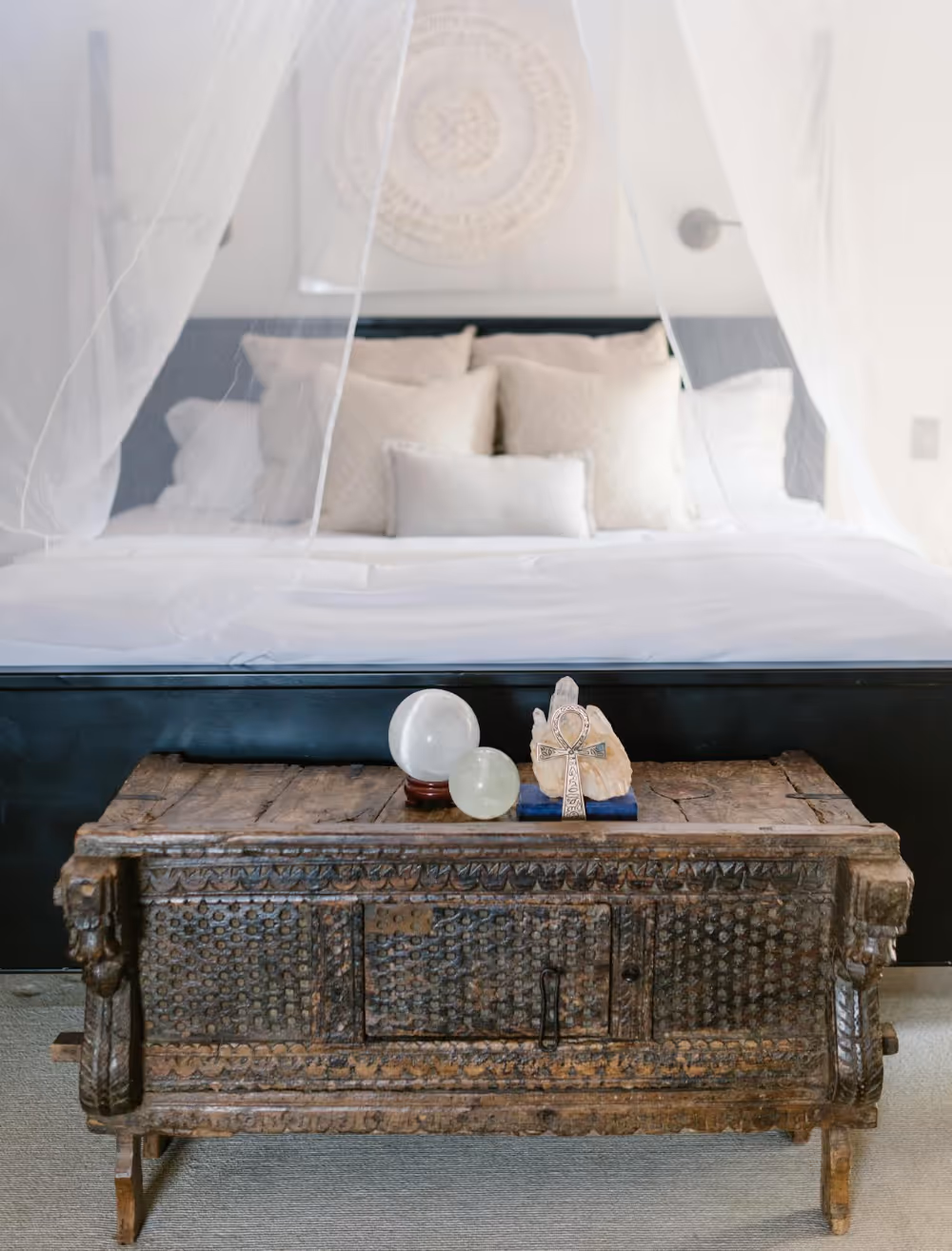 An antique blanket chest at the bottom of a bed with wispy voile curtains.